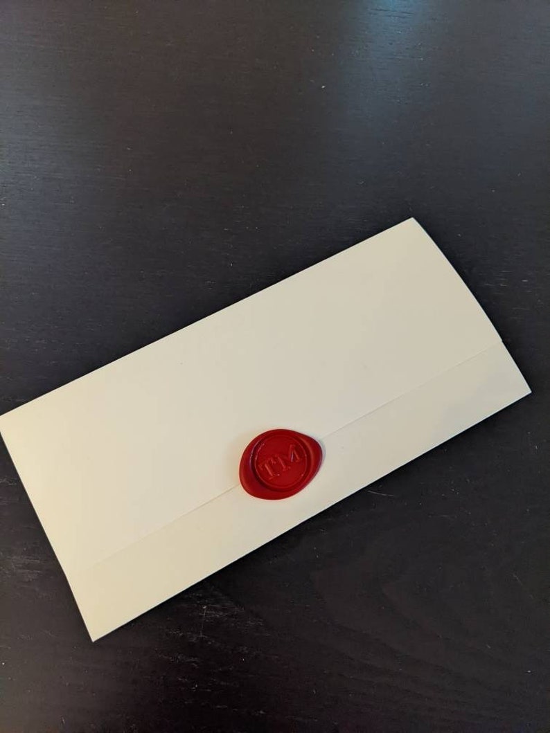 Puede incluir: Sobre de color marfil sellado con un sello de cera rojo. El sello presenta las iniciales "TM" en un dise&ntilde;o circular. El sobre est&aacute; colocado en diagonal sobre una superficie oscura.