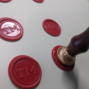 May include: Red wax seals with the letters "TM" imprinted on them. A wooden handle with a gold metal base is used to create the seals.