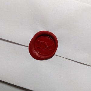 Puede incluir: Un sello de cera rojo con las letras "TM" en relieve sobre un sobre blanco. El sello circular tiene un acabado brillante. El sobre es blanco y tiene un dise&ntilde;o sencillo.