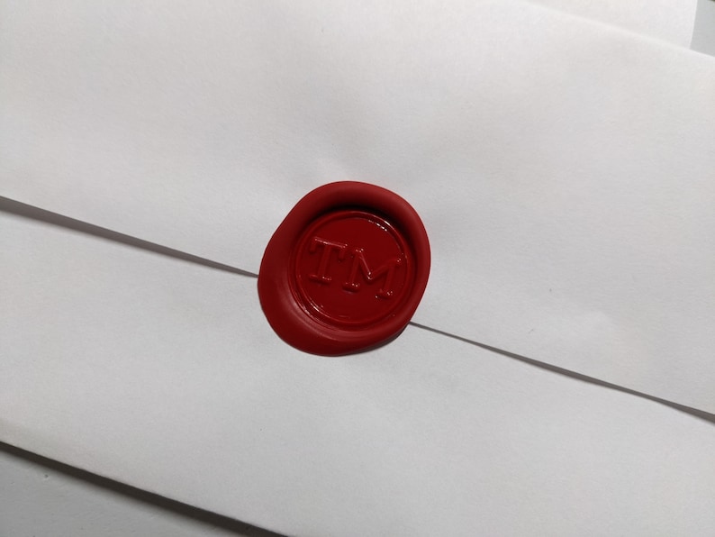 Puede incluir: Un sello de cera rojo con las letras "TM" en relieve sobre un sobre blanco. El sello circular tiene un acabado brillante. El sobre es blanco y tiene un dise&ntilde;o sencillo.