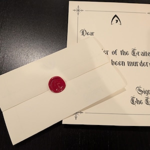 May include: An ivory-colored envelope sealed with a red wax stamp, next to a matching sheet of paper. The paper has gothic text that reads "Dear, er of the Traitors, been murdered. Signed, The Traitors."