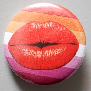 Puede incluir: Un botón redondo con un diseño a rayas rojo, blanco, naranja y rosa. Las rayas están en los colores de la bandera del orgullo lésbico. Una impresión de labios rojos está centrada en el botón.