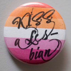 Könnte beinhalten: Ein runder Knopf mit dem Text "Kiss a Lesbian" in schwarzer Kursivschrift auf einem Hintergrund in den Farben der lesbischen Pride-Flagge: Orange, Weiß, Pink.