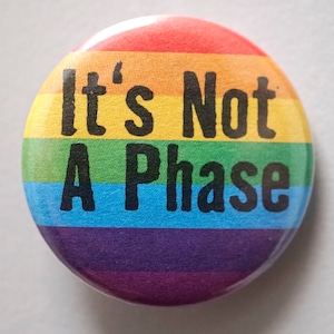 Op de afbeelding: Een ronde knop met de tekst "It's Not A Phase" in zwarte letters op een regenboogachtergrond.