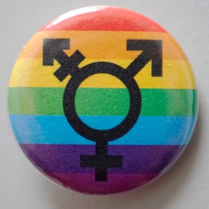 Könnte beinhalten: Ein regenbogenfarbener Knopf mit einem schwarzen Transgender-Symbol in der Mitte. Das Symbol ist ein Kreis mit einem nach rechts zeigenden Pfeil und einem Kreuz oben und einem Kreuz unten.