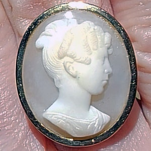 May include: A gold-rimmed oval cameo featuring a white cameo of a woman with her hair styled in a bun. The cameo is set in a gold frame.