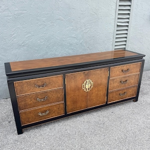 May include: A mid-century modern dresser with a black frame, burl wood drawer fronts, and a walnut top. The dresser has six drawers and two cabinet doors with a gold-colored geometric handle. The top measures approximately 72 inches long.