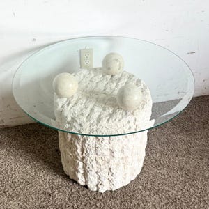 May include: A white, round, textured coffee table with a glass top. The table has four white, round orbs attached to the top.