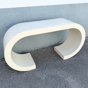May include: A cream-colored console table featuring a continuous, curved C-shaped design that forms both the top surface and the two side supports. The piece has a smooth, solid finish and a wide, open structure with rounded edges throughout.