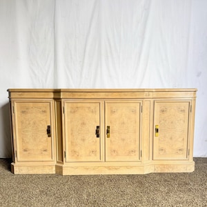 May include: A light-colored wooden cabinet with four doors and brass hardware. The cabinet has a decorative wood grain pattern and is set on a low base. The doors have rectangular panels and brass handles.