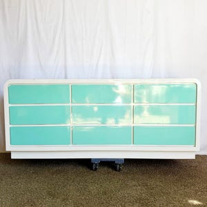May include: A white dresser with nine drawers, each painted a vibrant turquoise. The drawers are arranged in three rows of three, set within a white frame. The dresser sits on small wheels, suggesting mobility. The overall design is a blend of retro and modern aesthetics.