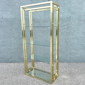 May include: A tall, gold-colored metal display shelf with three glass shelves. The open-frame design features a rectangular shape with a top and bottom, and vertical supports. The shelves are evenly spaced.