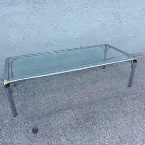 May include: A rectangular coffee table featuring a clear glass top supported by a polished chrome metal frame. The structure includes four straight legs connected by horizontal bars, with gold-toned metal accents at each corner joint.