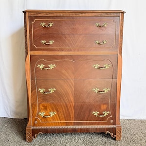 May include: A tall, antique wooden dresser with five drawers. The dresser is made of dark wood with a lighter wood trim and gold-colored handles. The top of the dresser has a decorative border. The drawers have a curved design.