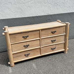 May include: A light-colored bamboo dresser with six drawers and a woven top. The dresser has dark metal handles and is made of natural bamboo. The design is a mix of rustic and tropical styles.