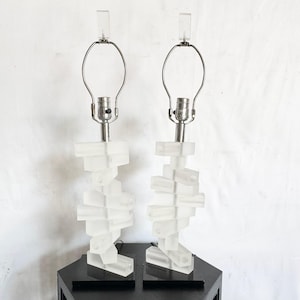 May include: Pair of frosted acrylic table lamps. Each lamp features a stacked, geometric base design with a silver-toned neck and a clear acrylic finial. The lamps are displayed on a black surface against a white background.