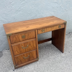 May include: A wooden desk with a warm brown finish. It features three drawers on the left side and a single drawer on the right. The desk has brass corner accents and drawer pulls, giving it a vintage look. The desk is sturdy and well-crafted.