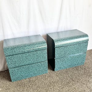 May include: Two matching teal-colored nightstands with a speckled granite-like finish. Each nightstand has a curved top and three drawers with horizontal grooves. The nightstands are placed side by side on a neutral-colored carpet.