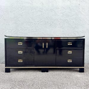 May include: A black dresser with six drawers and a cabinet door in the center. The dresser has gold-colored hardware and trim. The top of the dresser has a dark surface. The dresser is on a gray surface.