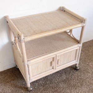 May include: A light beige rattan side table with two shelves and a cabinet with two doors. The top and middle shelf have a woven texture. The cabinet doors have vertical lines. The table has small wheels.