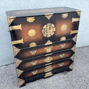 May include: A dark brown chest of drawers with brass hardware. The chest has four drawers and decorative brass corner brackets. The front of the chest has a gradient color from brown to black.