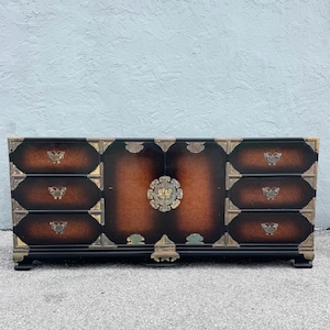 May include: A dark wood cabinet with a black base and brass hardware. The cabinet has drawers and doors with a central decorative element. The wood has a brown and black finish.