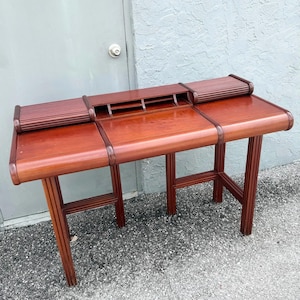 May include: A reddish-brown wooden desk featuring a smooth, flat work surface, two raised side sections with ribbed detailing, and a central storage compartment with three open slots. The desk is supported by four vertical, fluted legs.