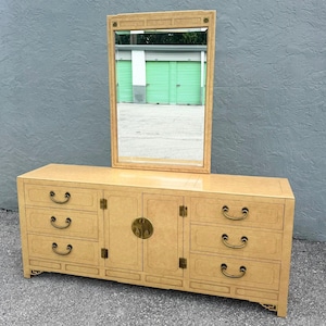 May include: A light-colored wooden dresser with a matching rectangular mirror. The dresser has multiple drawers and cabinet doors with brass hardware. The mirror is rectangular with a decorative frame. The furniture is placed against a gray wall.