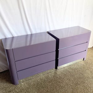 May include: Two matching lavender dressers with three drawers each. The dressers have a glossy finish and rounded edges. They are placed side by side on a neutral-colored carpet, against a white backdrop.