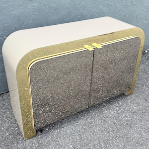 May include: A cream-colored cabinet with a mirrored front and gold trim. The cabinet has a rounded top and sides, with two doors that meet in the center. The gold trim frames the mirrored doors and edges.