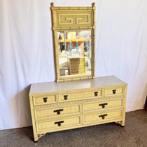 May include: A light-colored wooden dresser with a matching mirror. The dresser has seven drawers with dark hardware and decorative trim. The mirror is rectangular with a bamboo-style frame. The furniture is a light beige color.