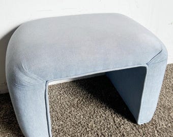 May include: A light blue upholstered ottoman with a curved, open design. The ottoman is sitting on a brown carpet.