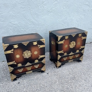 May include: Two matching black and brown wooden cabinets with gold-colored hardware. The cabinets have a rectangular shape with a two-door design and decorative floral accents. The hardware includes corner brackets and drawer pulls.