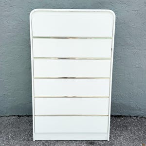 May include: A white six-drawer dresser with a rounded top and silver-colored horizontal accents. The dresser has a rectangular shape and is set against a gray textured wall.