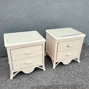 May include: Two matching off-white wicker nightstands with two drawers each. The nightstands have a woven design and glass tops. The drawers have small, light-colored wooden handles. The nightstands are on a gray surface.
