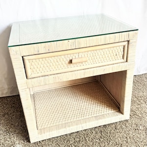May include: A light-colored woven nightstand with a glass top. The nightstand has a single drawer and an open shelf below. The drawer has a wooden handle. The nightstand is made of woven material.