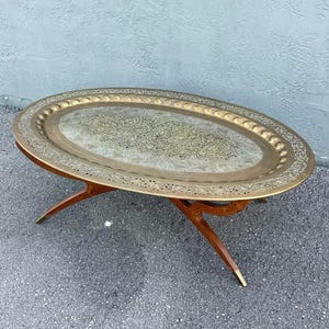 May include: An oval coffee table featuring a large, removable brass tray top with intricate etched floral patterns and a scalloped rim. The base is constructed from dark wood with four curved, intersecting legs that feature brass-capped feet.