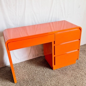 May include: A vibrant orange desk with a glossy finish. The desk features a rounded edge design and three drawers on the right side. The desk's dimensions are approximately 48 inches wide, 24 inches deep, and 30 inches high.