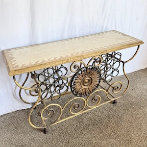 May include: A decorative console table with a rectangular top and ornate metalwork. The tabletop is light beige with a patterned border. The metal frame is gold with black accents, featuring scrollwork and a sunburst design. The table is approximately 48 inches long.