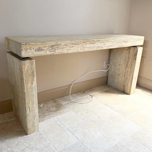 May include: A rectangular console table crafted from beige travertine stone with a porous, natural texture. The piece features a thick, flat top supported by two wide, vertical slab legs, creating a clean, monolithic structure with visible stone grain.