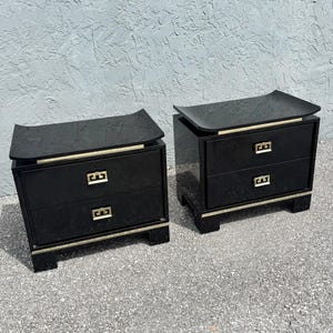 May include: Pair of black nightstands with two drawers each. The nightstands have a curved top and gold-colored trim. The drawer pulls are gold-colored. The nightstands are on a gray surface.