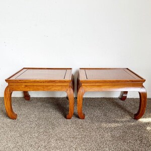 May include: Two matching wooden side tables. Each table has a rectangular top with a dark border and curved legs. The tables are made of a light brown wood and sit on a neutral-colored carpet.