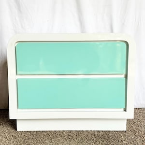 May include: A white two-drawer nightstand with rounded corners and a mint green drawer front. The nightstand has a rectangular base and is set against a white backdrop.