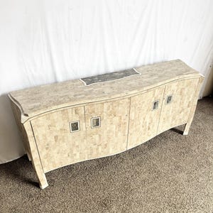 May include: A long, curved cabinet with a mosaic-like pattern in shades of beige and cream. The cabinet has four doors with square silver handles and a dark gray accent on the top. The legs are white.
