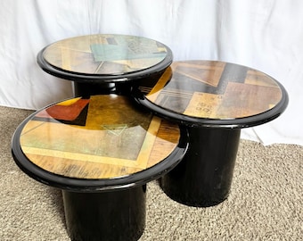 May include: A set of three nesting tables with cylindrical black bases and circular tops. Each tabletop features a glossy, multi-colored geometric pattern with shades of brown, orange, yellow, and green, finished with a smooth black rim.