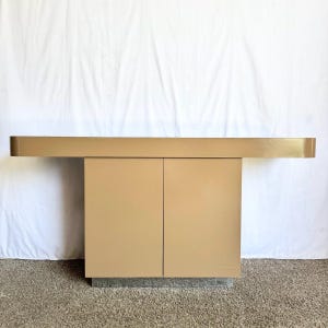 May include: A tan-colored cabinet with two doors and a long, horizontal top. The cabinet has a rectangular base and a simple, modern design. The top extends beyond the cabinet's base on both sides.