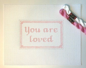 May include: A white canvas with a pink frame and the words "You are loved" in pink lettering. Two skeins of pink and white embroidery floss are included.