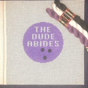 May include: Purple canvas with a white circle that says "The Dude Abides" and three black bowling pins. The canvas is accompanied by three skeins of embroidery floss: black, white, and purple.