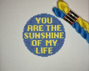 May include: A blue and yellow needlepoint canvas with the text "You are the sunshine of my life" in a circle. Two skeins of embroidery floss, one yellow and one blue, are on the canvas.