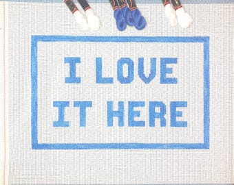 May include: White canvas with a blue border and the words "I LOVE IT HERE" stitched in blue thread.